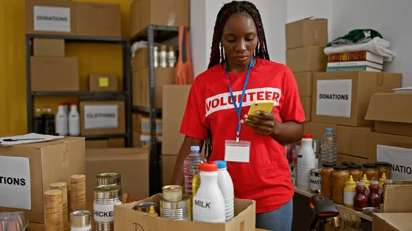 Dedicated african american woman volunteer diligently checking donated food products using smartphone at charity warehouse