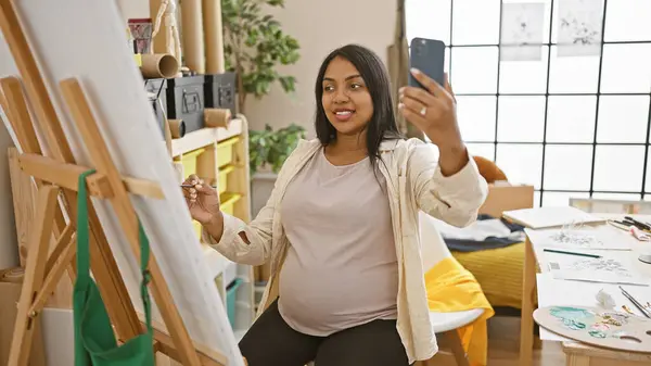 Expecting young latina artist, joyfully smiling as she draws her motherhood journey in her art studio, while engaging in a video call for her university painting lesson.