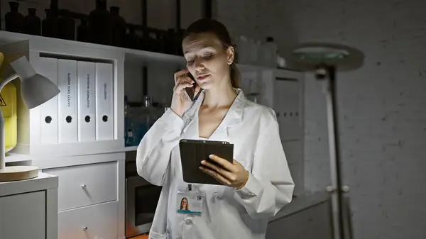 A young woman in a lab coat multitasking in a pharmacy with a phone and tablet.