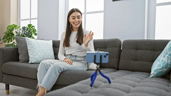 Smiling young hispanic woman recording a video on phone with a tripod in a cozy living room setting.