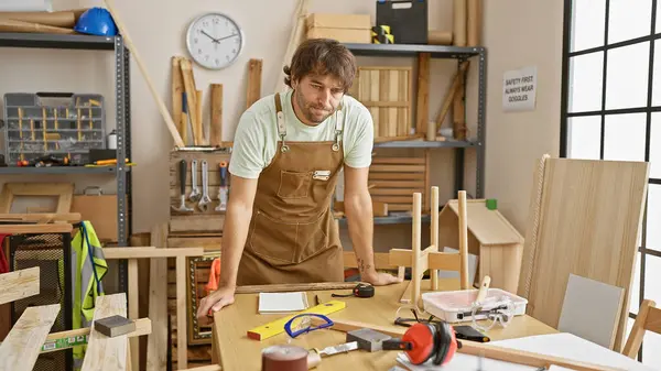 一个留着胡子的年轻人在一个木匠工作室里 四周都是工具和木制品 传达着手艺 — 图库照片