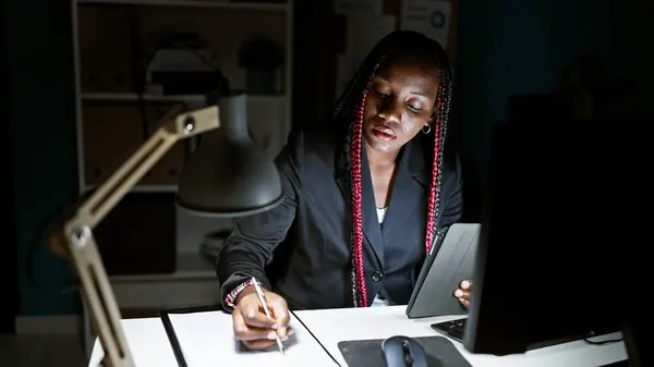 Serious african american woman boss takes business notes on touchpad, focused in the office night light, with dark glasses on, beautifully braided hair, a real professional
