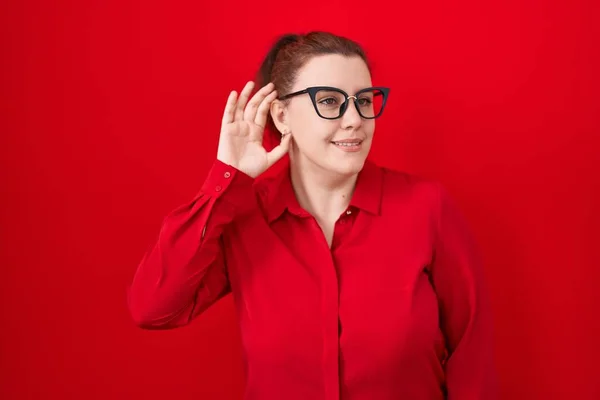 Young hispanic woman with red hair standing over red background smiling with hand over ear listening an hearing to rumor or gossip. deafness concept.