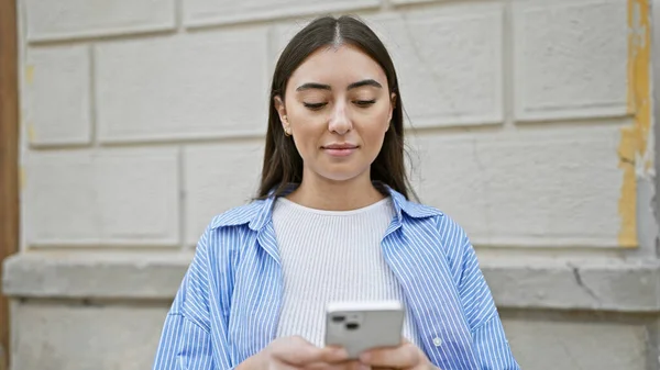 A young hispanic woman in casual attire uses a smartphone on a city street, embodying urban lifestyle.