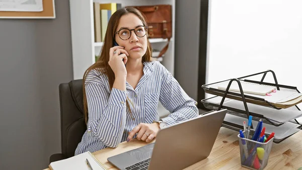 メガネの思慮深い女性は 現代のオフィスルームのラップトップと机に座り プロの職場環境を描いています — ストック写真