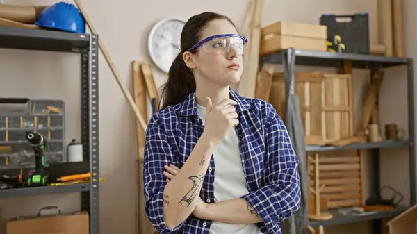 一位头脑清醒 戴着安全眼镜的年轻女子站在一个被木工工具环绕的车间里 — 图库照片