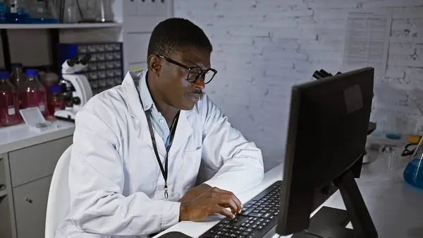 African man in lab coat working on computer in a laboratory setting with scientific equipment