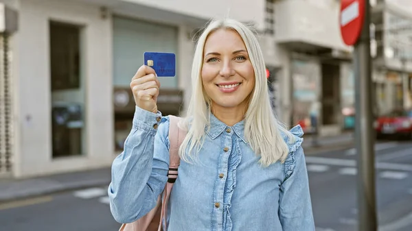 Confident young blonde woman joyfully holding a credit card, smiling radiantly in the sunlit city street. her casual fashion exudes a lifestyle of financial freedom and effortless charm.