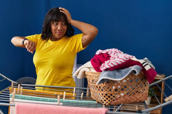 Young beautiful latin woman looking clock hanging clothes on clothesline at laundry room