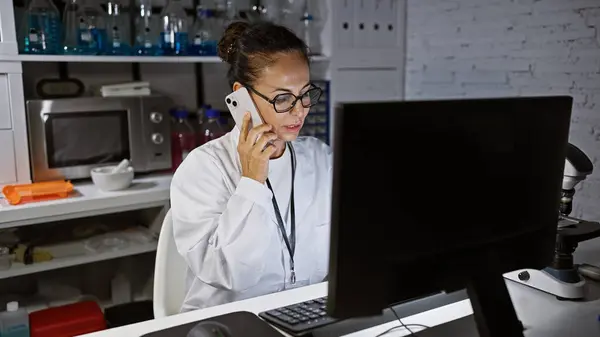 A mature hispanic woman in a lab coat talks on the phone while working on a computer in a laboratory setting.