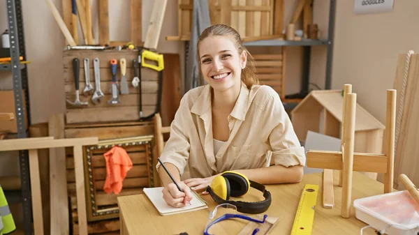 一位面带微笑的年轻女子在一个被工具和木头环绕的木工车间里画草图 — 图库照片