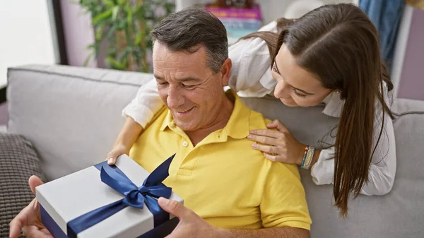 At home, a surprised father and daughter smiling together, sitting on the sofa with a gift in a loving hispanic familys relaxing living room.