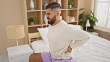 A bearded man grimaces with back pain sitting on a white bed inside a bedroom, showing a cozy, modern home interior.