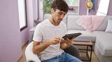 Soğukkanlı, genç İspanyol bir adam, gülümsüyor, rahat ev koltuğunda dinleniyor, dokunmatik dokunmatik bilgisayarındaki rahatlatıcı bir videoya dalmış, rahat kulaklıktaki sesten keyif alıyor.