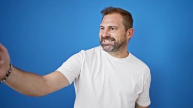 Beyaz gömlekli olgun sakallı bir adam özçekim yapıyor ve mavi arka planda yüz ifadeleri yapıyor..
