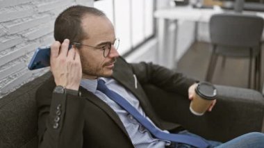 Sakallı, gözlüklü ve takım elbiseli profesyonel bir kel modern bir ofiste elinde kahve tutarken akıllı bir telefonla konuşuyor..