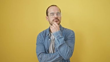 Sarı bir duvarın önünde kot pantolon giymiş, sakallı ve gözlüklü kendinden emin kel bir adam.