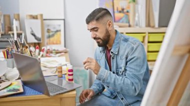Sakallı adam resim atölyesinde dizüstü bilgisayar ve sehpayla boya inceliyor..