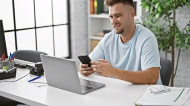 Kapalı ofis ortamında akıllı telefon ve dizüstü bilgisayar kullanan, sakallı, genç İspanyol bir adam..