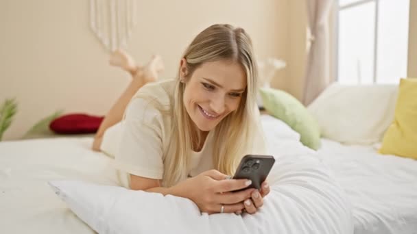 Έκπληκτη Όμορφη Νεαρή Ξανθιά Γυναίκα Ξαπλωμένη Στο Κρεβάτι Χρησιμοποιώντας Smartphone — Αρχείο Βίντεο