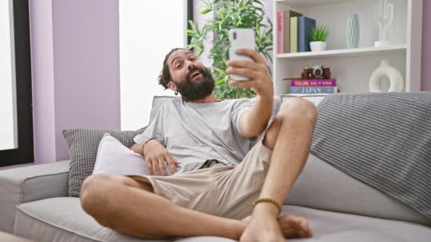 一个快乐的大胡子男人躺在家里的沙发上 一边拿着手机一边高兴地做着一个和平的手势 — 图库视频影像