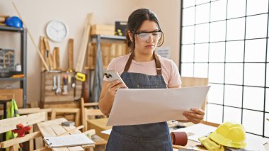 Odaklanmış bir İspanyol kadın elinde akıllı bir telefonla güneşli bir marangoz atölyesinin planını inceliyor..