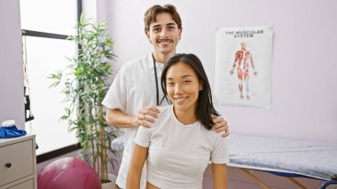 Rehabilitasyon kliniğinde arka planda anatomik posteri olan bir kadın hastayla gülümseyen erkek terapist..