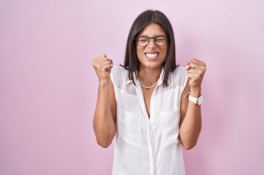 Pembe arka planda duran esmer, başarı için heyecanlanan gözlüklü, kolları havada ve gözleri kapalı zaferi kutlayan genç bir kadın. Kazanan konsept. 