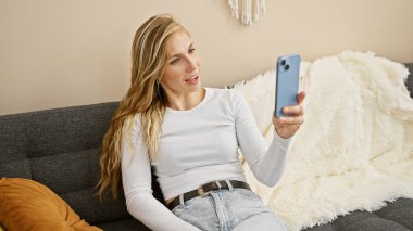 Çekici genç kadın rahat oturma odasında akıllı telefon kullanıyor, evde teknoloji kullanımı tasvir ediyor..