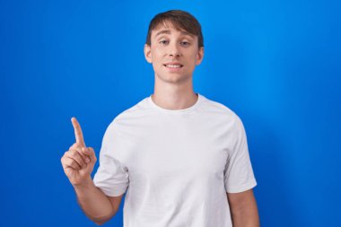 Mavi arka planda duran beyaz tenli sarışın adam bir numaralı parmağı gösterip işaret ederken kendinden emin ve mutlu gülümsüyor.. 