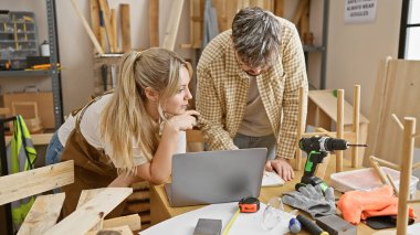 Bir erkek ve kadın, aletler ve tamamlanmamış mobilyalarla çevrili bir marangozluk stüdyosunda iş birliği yapıyor..