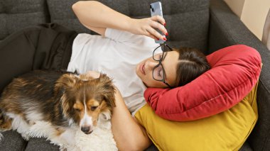 Genç bir İspanyol kadın, köpeği ile kanepede rahatlar, rahat bir evde akıllı telefonunu karıştırır.