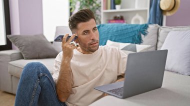 Evde yerde oturmuş, akıllı telefondan sesli mesaj dinlerken dizüstü bilgisayar kullanan odaklanmış genç bir İspanyol.