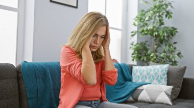 Sıkıntılı, orta yaşlı, sarışın bir kadın, evin içinde gri bir koltukta oturuyor. Endişelerini ya da baş ağrılarını bir oturma odasına yansıtıyor..