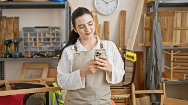 Ahşap atölyesinde beyaz gömlek ve önlük giyerken akıllı telefon kullanan genç bir İspanyol kadın..