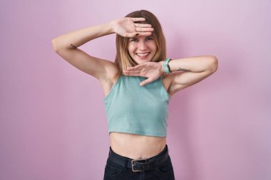 Blonde caucasian woman standing over pink background smiling cheerful playing peek a boo with hands showing face. surprised and exited 