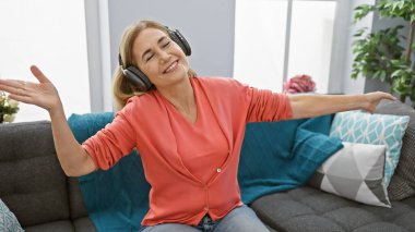 Orta yaşlı sarışın kadın evde kulaklıklarla neşeyle dans ediyor..