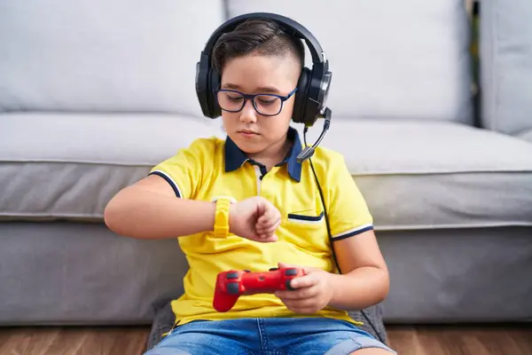 若いヒスパニックの子供は 腕時計の時間をチェックするヘッドフォンを身に着けているコントローラーを保持するビデオゲームをプレイ リラックスして自信を持って — ストック写真