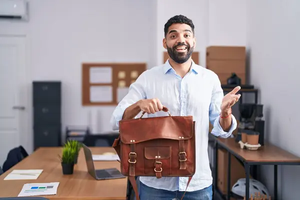 Hispanic man with beard working at the office holding briefcase celebrating achievement with happy smile and winner expression with raised hand