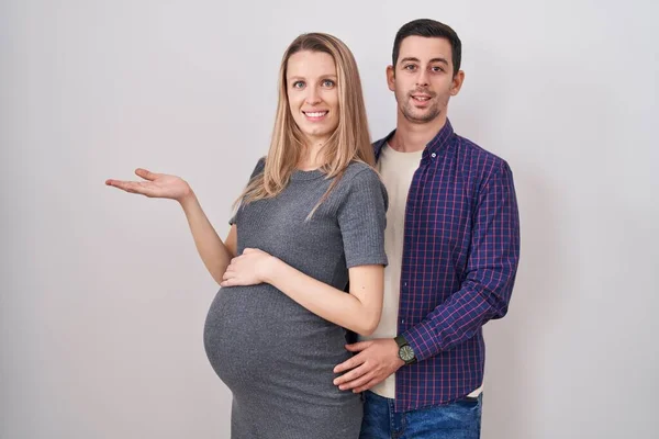 若いカップルは 赤ちゃんが白い背景の上に立っていることを期待して コピースペースを示す手を開いた手のひらを脇に置いて 興奮する笑顔の広告を提示 — ストック写真