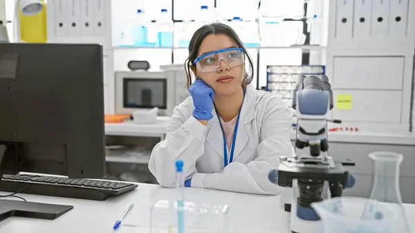 Contemplative Hispanic Woman Scientist Lab Coat Safety Glasses Sits Laboratory — Stock Photo, Image