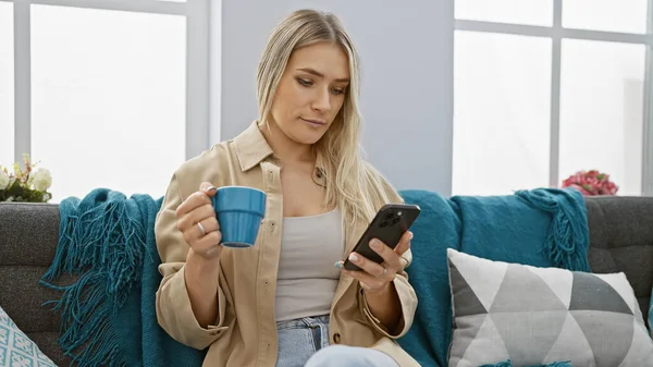 Young, attractive blonde woman lost in thought, relaxing at home with a cup of coffee. concentrated on her smartphone, typing a serious message. indoor portrait, sitting on the sofa.