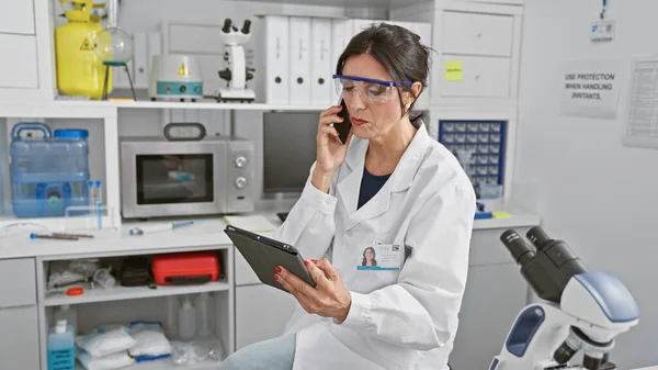Hispanic woman scientist in lab coat using tablet and talking on phone in laboratory
