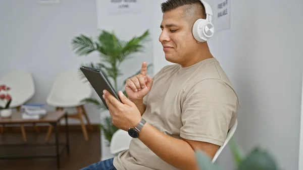 冷冰冰的年轻拉丁裔男人喜欢用耳机唱着动听的歌曲 舒服地坐在候车室里 自信地在触摸板上对着摄像机微笑 — 图库照片