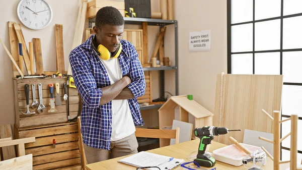 Handsome black man with crossed arms standing thoughtfully in a carpentry workshop with tools and wood around.