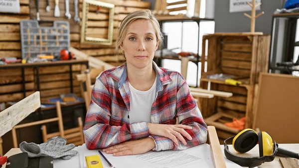 A confident woman carpenter sits in a workshop surrounded by tools and wood planks.
