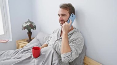 Orta yaşlı bir adam yatakta telefonla konuşuyor, elinde kırmızı bir kupa, rahatlamış, yatak odasında..