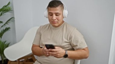 Akıllı telefon ve kulaklık kullanan genç latin adam bekleme odasında sandalyede oturuyor.