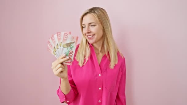 楽観的な若いブロンドの女性は喜んで新しいゼーランドドルを保持し 自信を持って手と指で側面を指し 孤立したピンクの背景に微笑みます — ストック動画