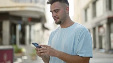 Sakallı genç bir İspanyol adam güneşli bir şehir sokağında akıllı telefon kullanırken gülümsüyor..